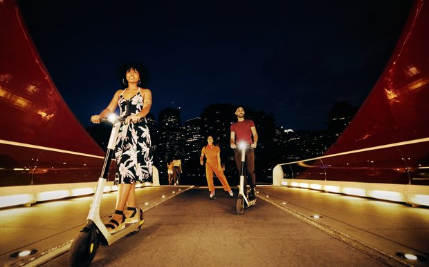 Two friends on electric scooters with lights, closely followed by a third person on roller skates, crossing the Peace Bridge at night.