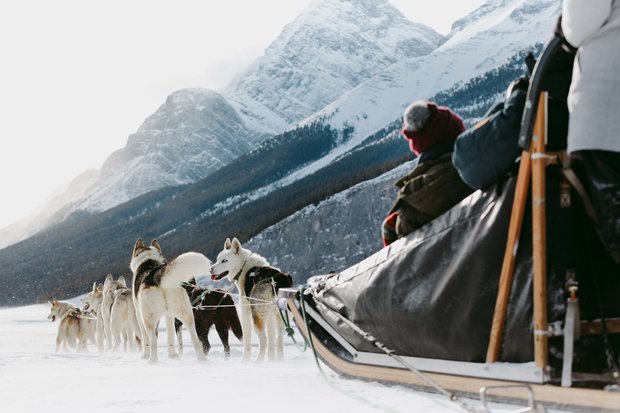 A ground level perspective as a dog sledding tour crosses a frozen lake with mountains in the background.