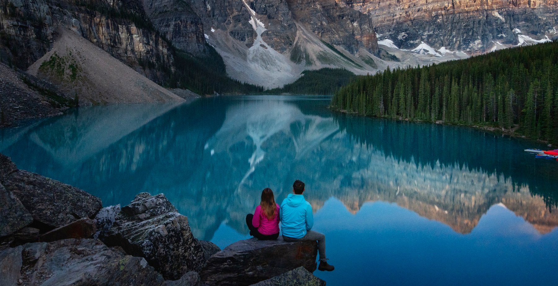 Couple sitting on a rock at the edge of Moraine Lake enjoying the mountain sunrise.