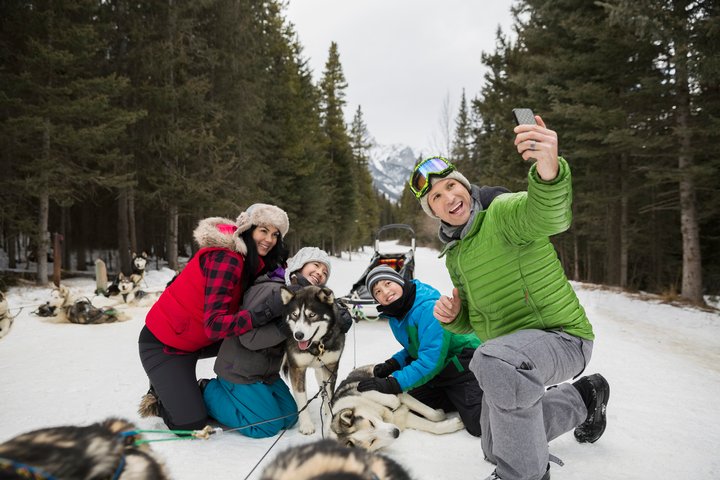 Family taking a smiling to take a selfie photo with the sled dogs in a snow covered forest.