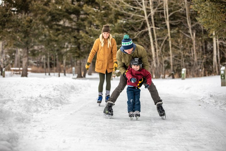 A family ice skates, Dad holding up his small child as Mom skates behind, at an outdoor ice path in Cypress Hills Provincial Park.