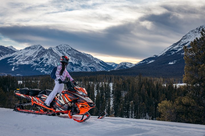 A woman on a snowmobile overlooking trees and snow-covered mountains.