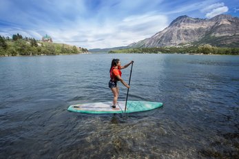 A paddleboarder paddles Middle Waterton Lake with the Prince of Wales Hotel on a hill and mountains in the distance.