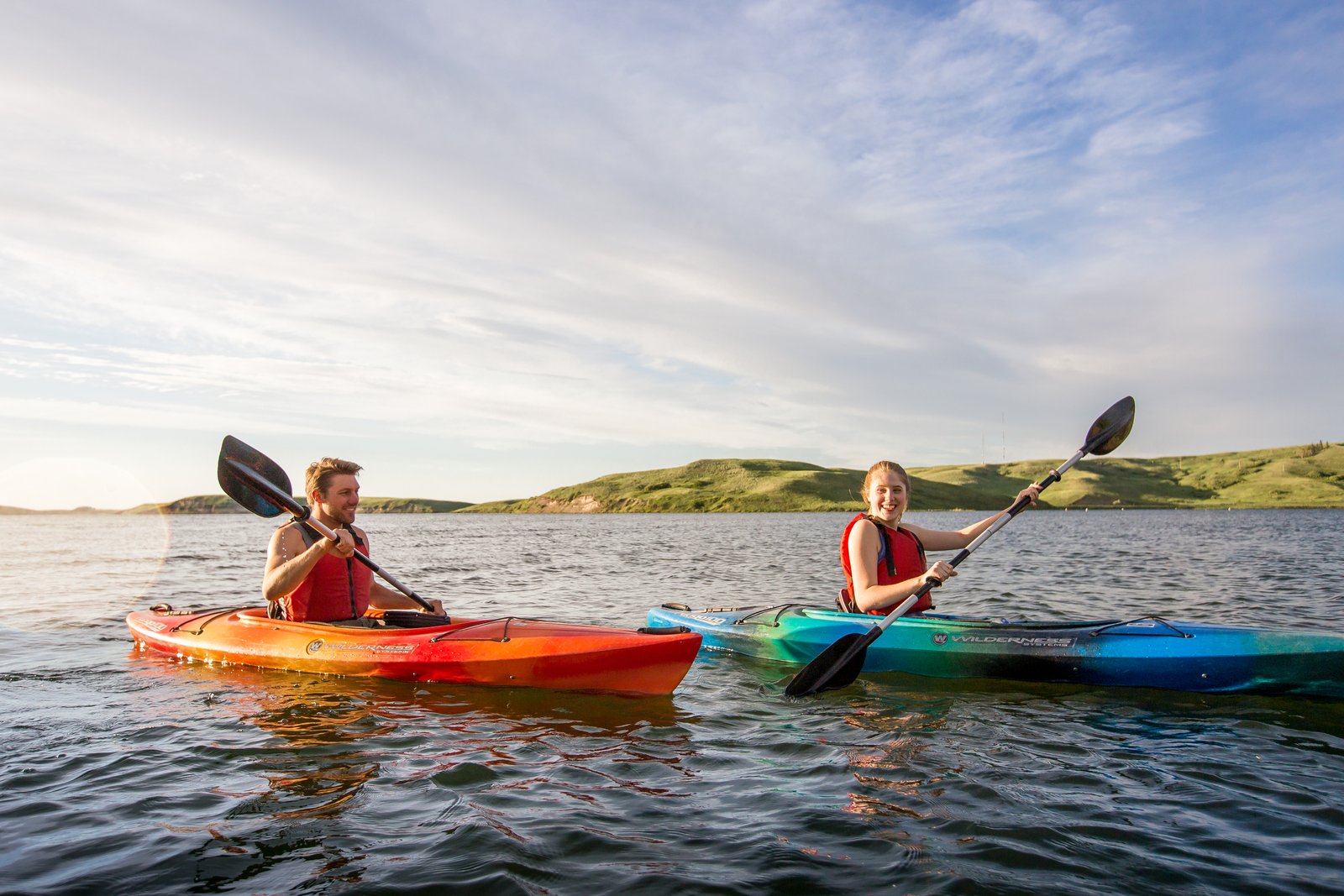 Couple kayaking on Elkwater Lake with green rolling hills in the background in Cypress Hills Interprovincial Park.