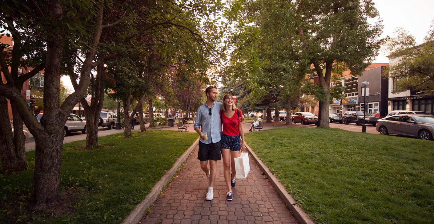 Couple walks along a tree-lined path with a shopping bag and a coffee.