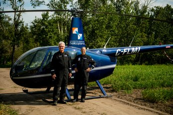 Two men in pilot jumpsuits standing in front of a helicopter on a remote dirt road