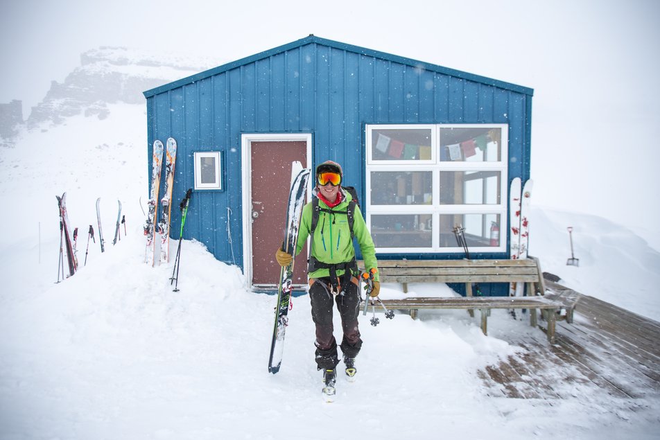 A man dressed in winter clothing and holding a pair of skies stands in front of a small lodge on the top of a snowy mountain.