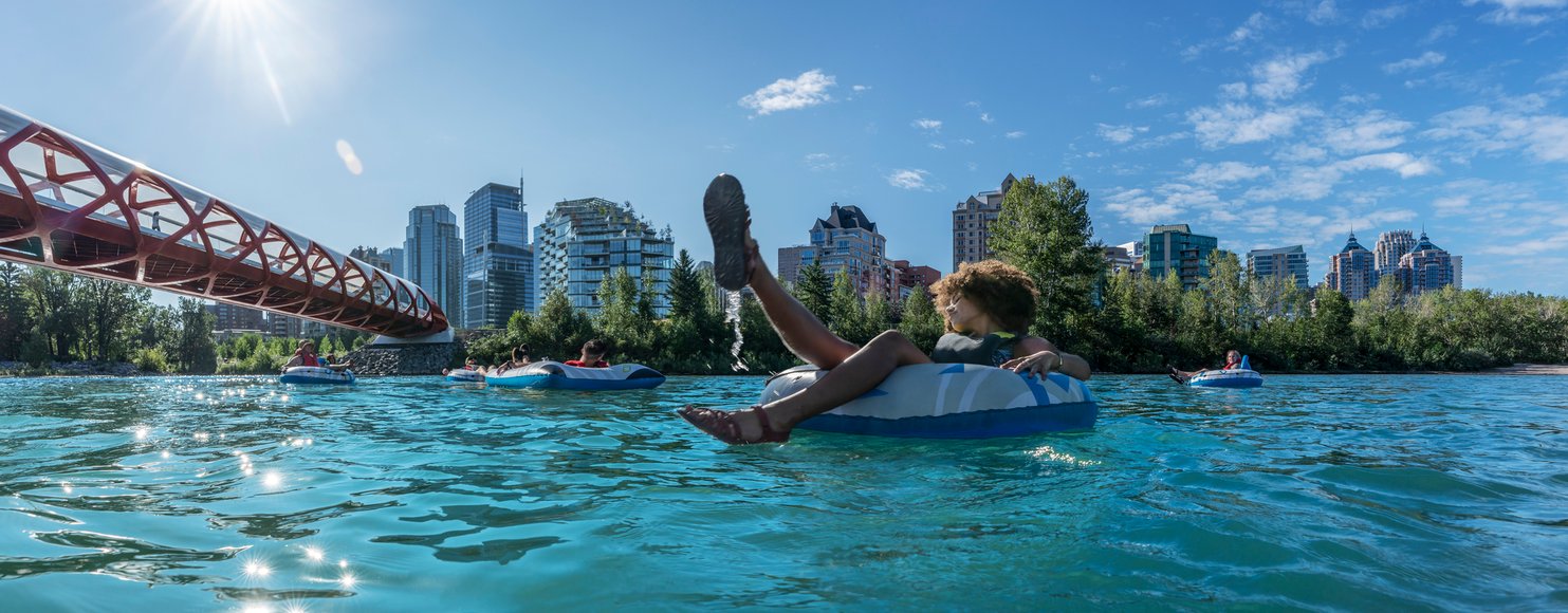 Young friends in swimsuits & buoyancy aids float leisurely on inner tubes down the Bow River in Calgary. Tree-lined banks & distant cityscape provide scenic backdrop as they approach Peace Bridge on sunny day, creating a peaceful & carefree atmosphere.