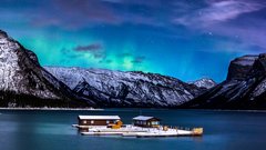 Northern lights in the winter nights sky above a floating dock on a lake with mountains in the background.