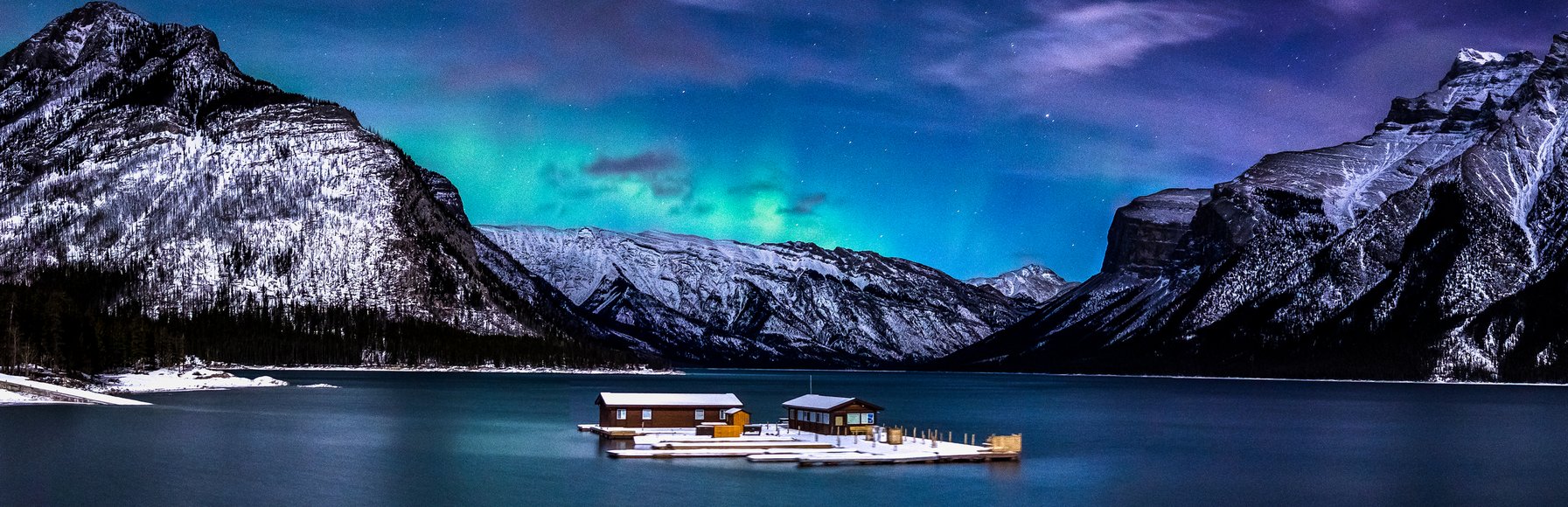 Northern lights in the winter nights sky above a floating dock on a lake with mountains in the background.