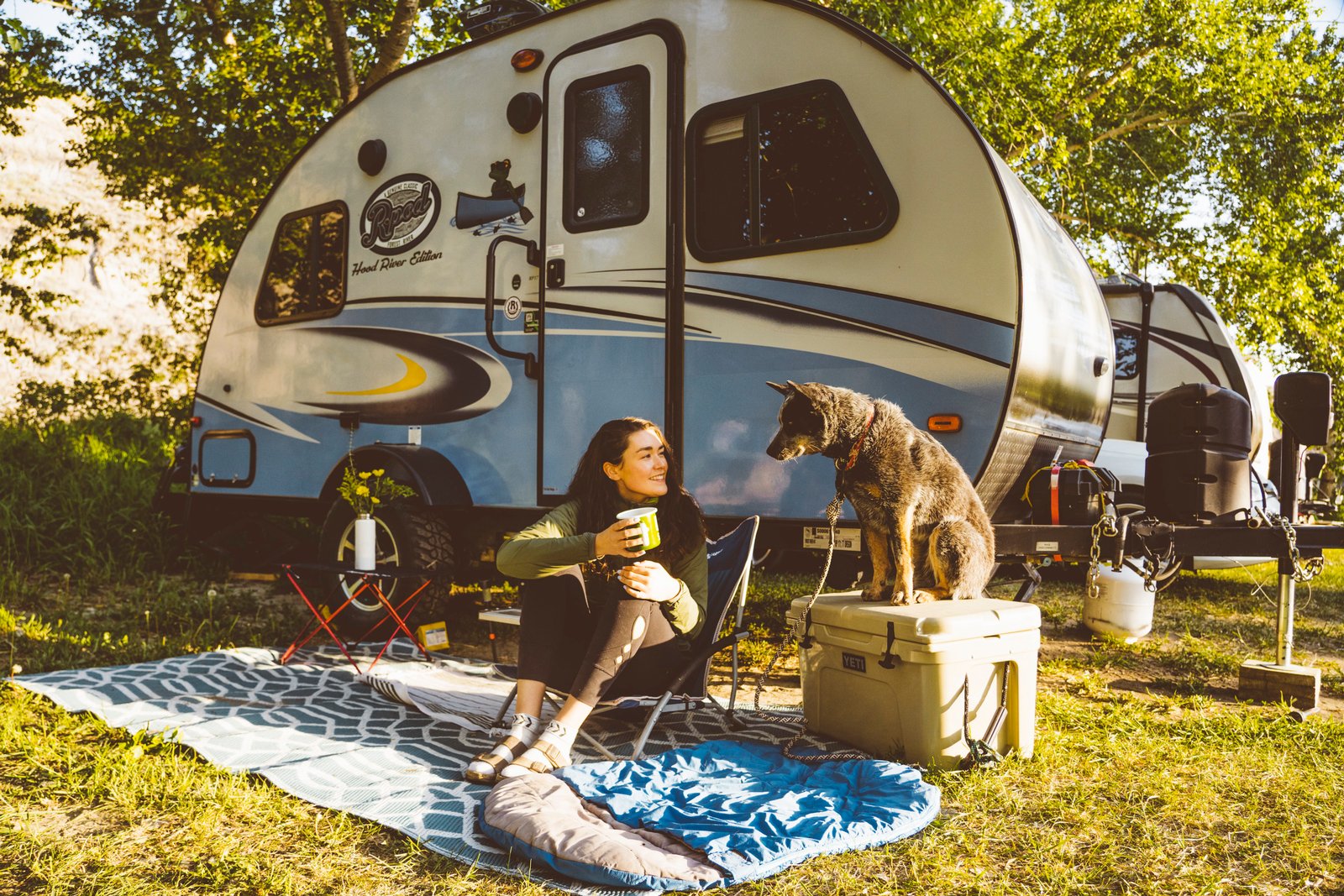 A woman camping with her dog sitting on the ground in front of their RV.