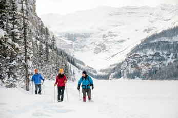 Travellers dressed in bright winter gear, snowshoe at the edge of an alpine forest with a snow-covered mountain in the background.
