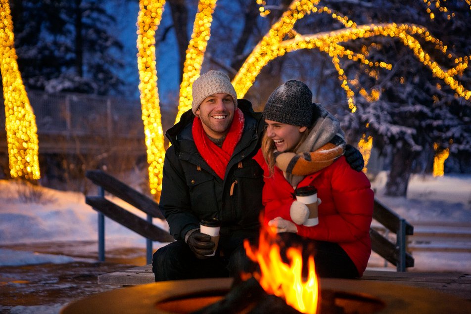 A couple enjoying hot chocolate in front of an outdoor fire pit at night.