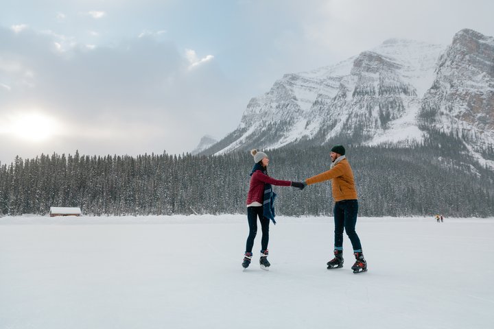 A couple holding hands while ice skating on a frozen lake with mountains and trees in the background.