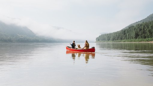 Couple / man and woman and dog paddling on a lake in a red canoe with misty forest and mountain scene in the background