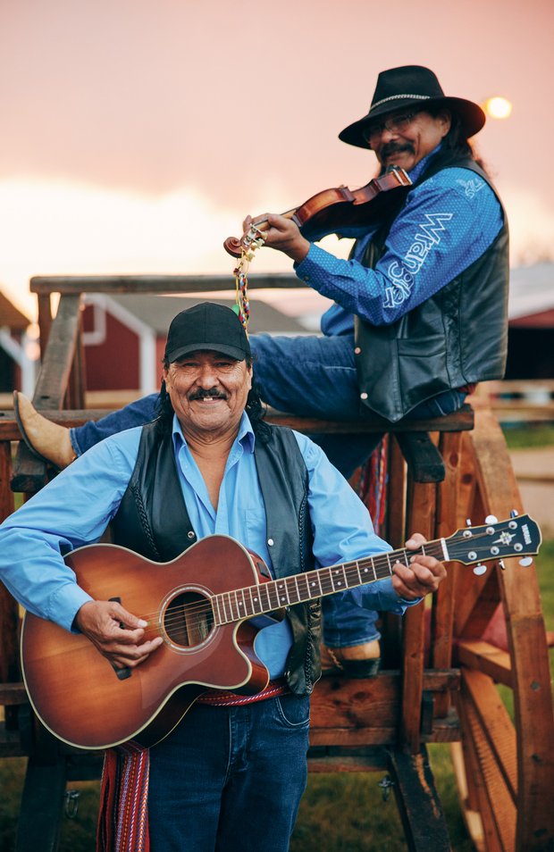 Two male Métis musicians smiling while sitting on a large wooden wagon with their instruments.