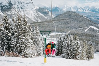 A couple ride a chairlift at Mt. Norquay on an idyllic winter day, with snow-dusted trees and mountains surrounding them.