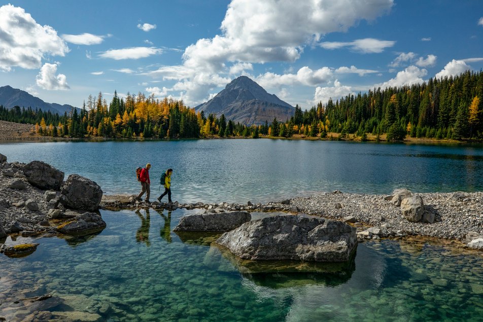 A couple hikes across rocks along a lake with larch trees and mountains in the background.