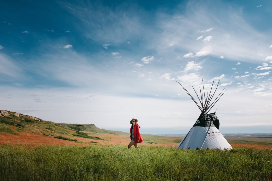 A woman walks beside a tipi in an open field with hills in the background.