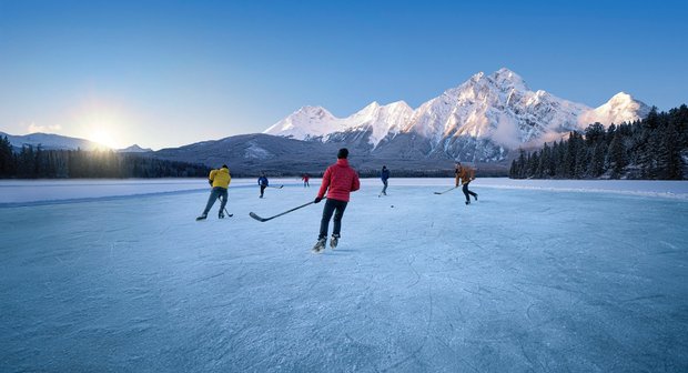 A group of people playing a game of pond hockey as the sun goes down, mountains in the background, on Pyramid Lake in Jasper National Park.