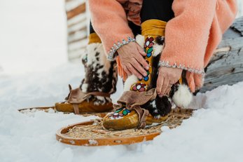 An Indigenous woman in a peach-coloured traditional coat attaches snowshoes to moccasins.