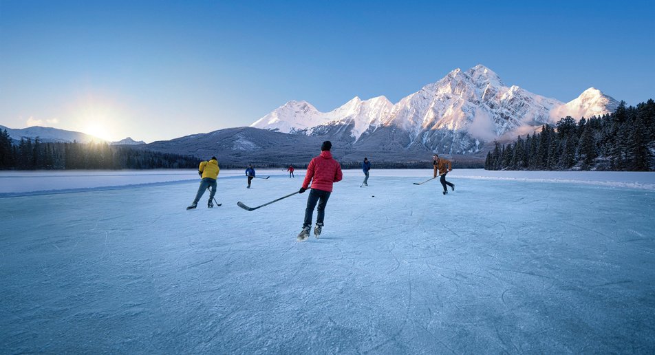 A group of people playing a game of ice hockey on a frozen lake as the sun goes down with mountains in the background.