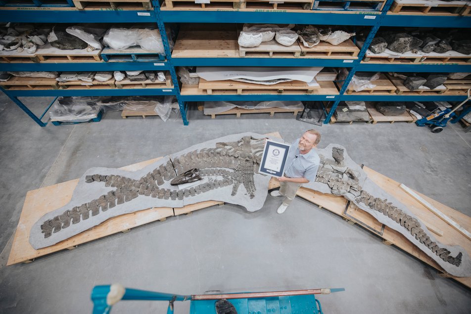 A man stands beside the fossil of a dinosaur holding a framed Guinness World Records certificate