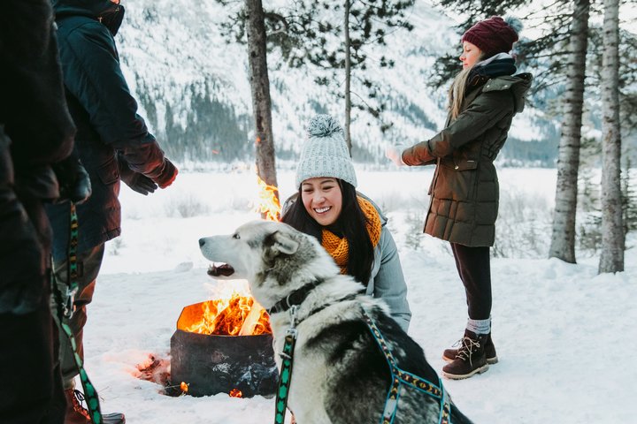 Two people standing around an outdoor fire while one smiles and pets a Husky after a dog sled adventure in the winter mountains.