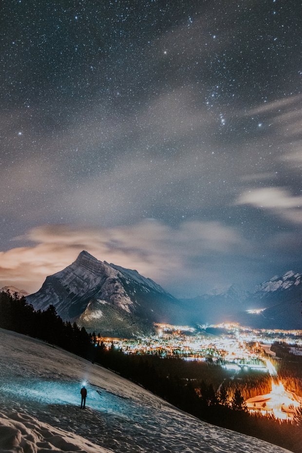 Person standing in the snow on Mount Norquay with views of Banff town lights under a starry night sky