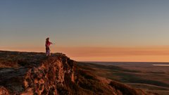 An indigenous man standing on the edge of Head-Smashed-In Buffalo Jump with the sun setting on the horizon.