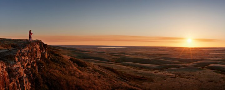 An indigenous man standing on the edge of Head-Smashed-In Buffalo Jump with the sun setting on the horizon.