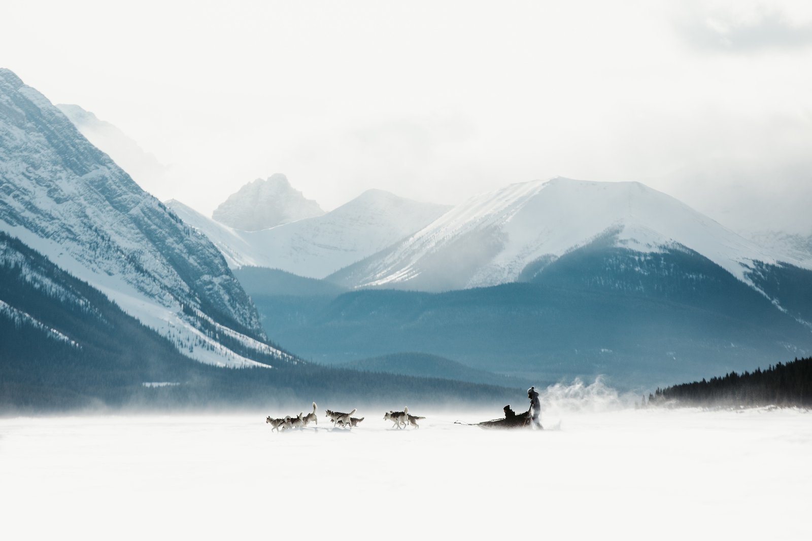 A dog sled crosses a frozen lake causing snow to spray up behind the riders with mountains in the background on a wintery day.