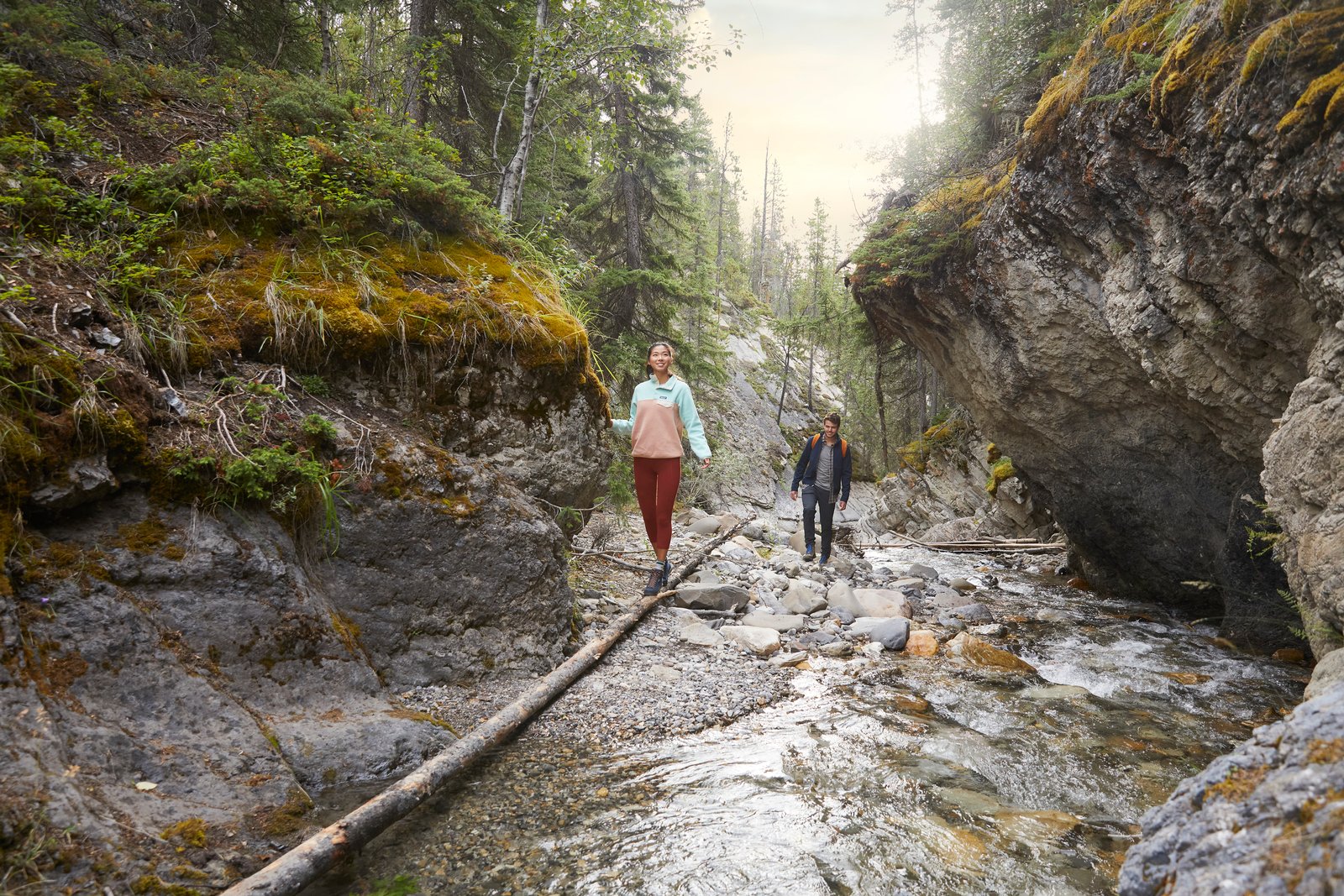 A woman balancing on a fallen down tree trunk with a man behind her standing on rocks as they are on a hike together admiring Whitegoat Falls in Nordegg.