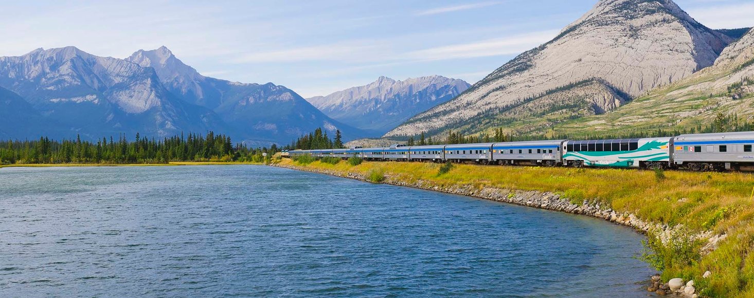 VIA Rail train travelling along a pristine lake on a sunny day as it enters the Rock Mountains towards Jasper.