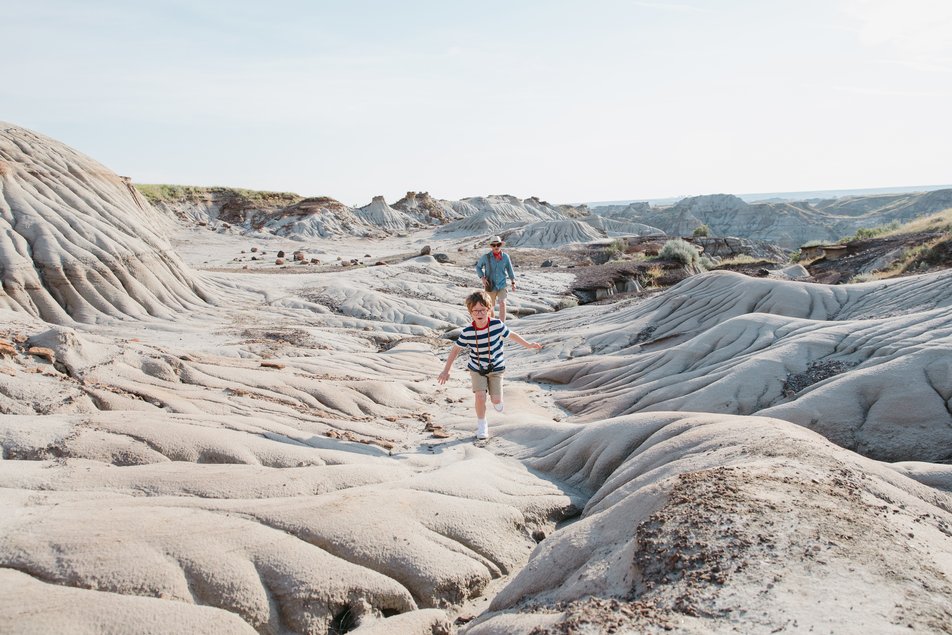 A little boy runs along rock coulees in the Canadian badlands, with his parent standing behind him.