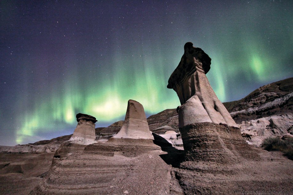 The green aurora borealis lights up a starry night sky. In the foreground, hoodoo rock formations loom.