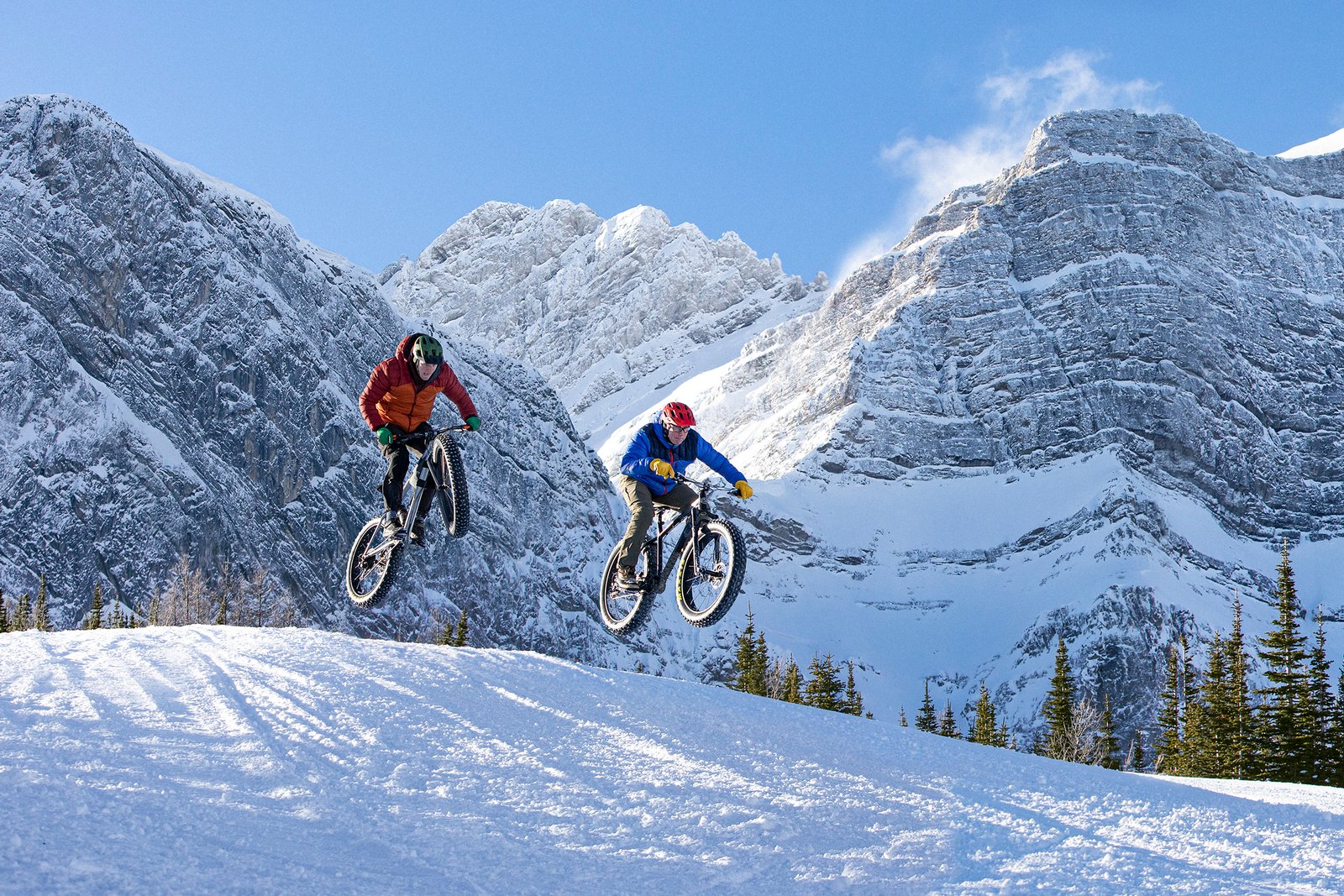 Two people fatbiking and getting air, on a brilliant winter's day, mountains in the background in Kananaskis Country.