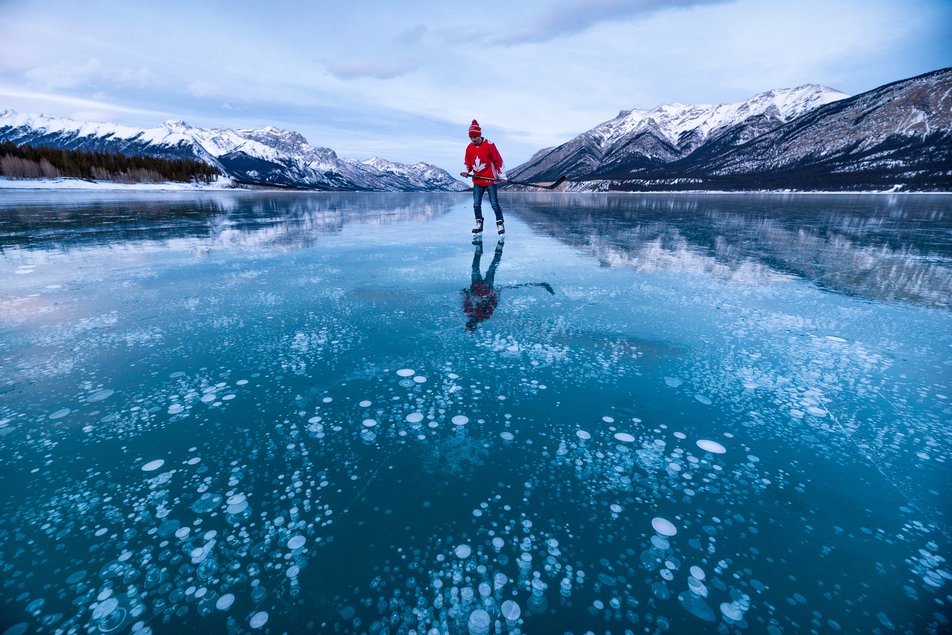 A person ice skating on a frozen lake with bubbles underneath the surface and mountain views in the background.