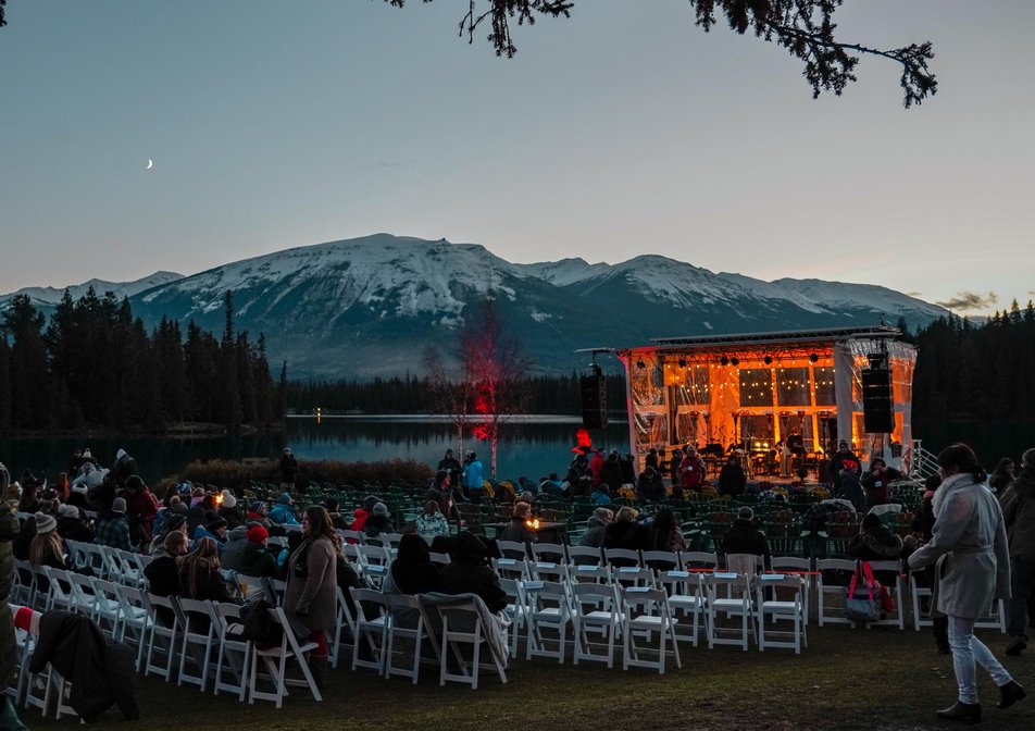 A crowd gathers in white chairs facing a lit stage with the Rocky Mountains in the background.