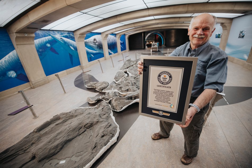 A man stands in the Royal Tyrrell Museum beside a Shonisaurus skeleton, holding a framed Guinness World Records certificate.