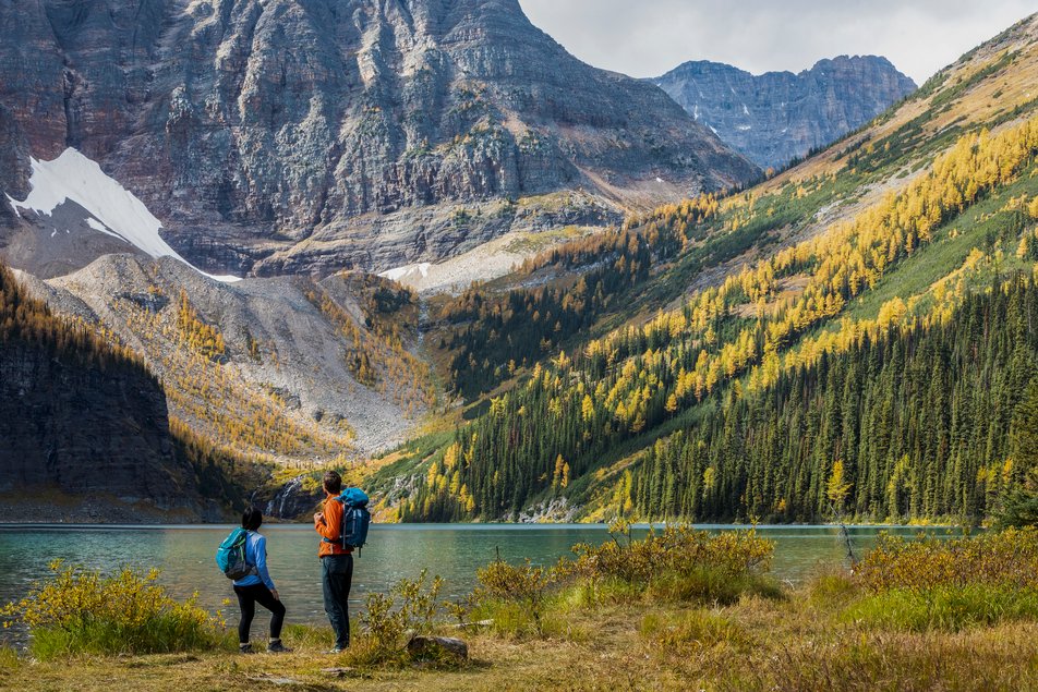 Two people stand by a lake with larch trees and mountains around them.