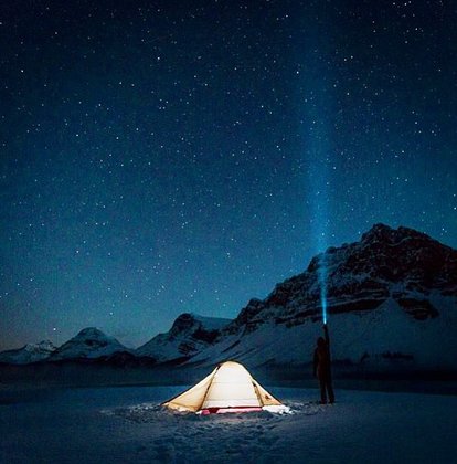A person beside a tent at night, shining a headlamp into the dark starry sky while winter camping in the mountains.