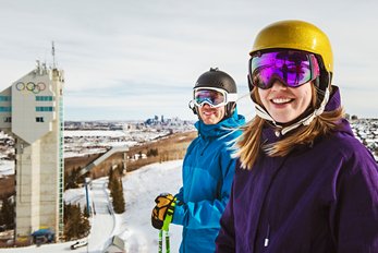A man and woman at the top of a run at WinSport in Calgary, with the tower displaying Olympic rings in the background.