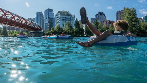 Young friends in swimsuits & buoyancy aids float leisurely on inner tubes down the Bow River in Calgary. Tree-lined banks & distant cityscape provide scenic backdrop as they approach Peace Bridge on sunny day, creating a peaceful & carefree atmosphere.