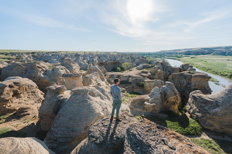 A man stands on top of a rock looking out at some hoodoos beside a river in the Canadian badlands.
