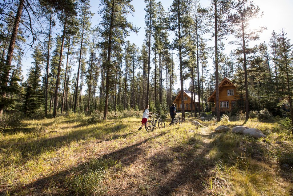 A young couple walking their bikes to a log cabin in the trees at Alpine Village in Jasper National Park.