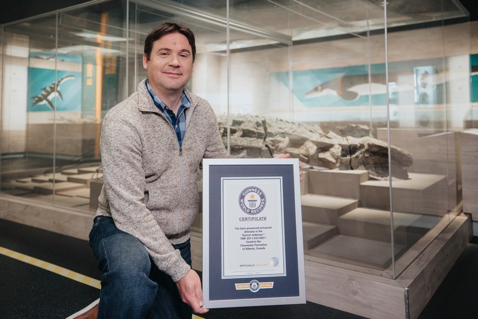 A man holding a framed Guinness World Records certificate stands in front of a dinosaur fossil.