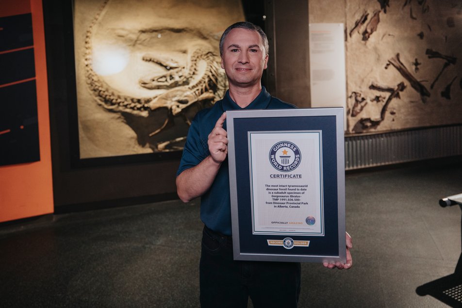 A man holding a framed Guinness World Records certificate stands in front of a dinosaur fossil.