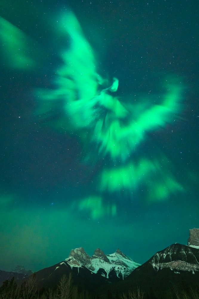 A green aurora borealis resembling an eagle hangs in the night sky above the Three Sisters mountains.
