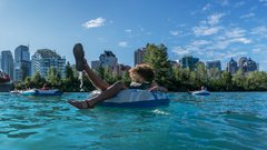 oung friends in swimsuits & buoyancy aids float leisurely on inner tubes down the Bow River in Calgary. Tree-lined banks & distant cityscape provide scenic backdrop as they approach Peace Bridge on sunny day, creating a peaceful & carefree atmosphere.